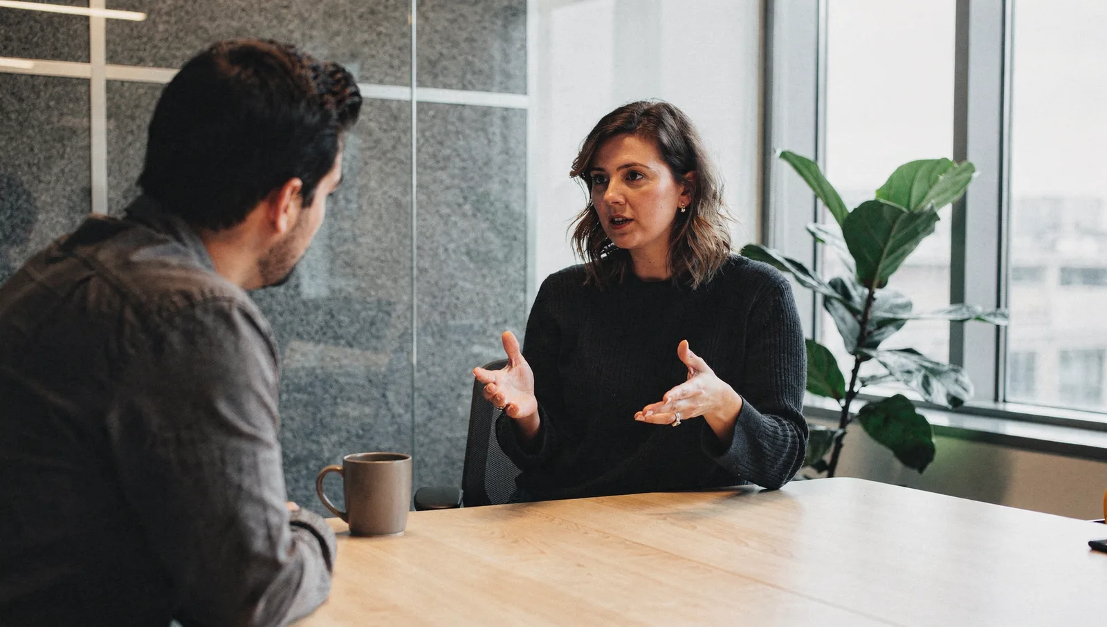 A woman gestures while speaking to a colleague at a conference table in a modern, sunlit office breakout room.